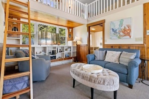A spacious living room with a high ceiling, a blue sofa, a wooden ladder, and a bookshelf filled with books and decorative items. - Little White Caps, Quaint Maine Cottage (Tenants Harbor)
