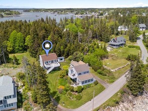 A residential area with houses surrounded by dense forest, a road, and a body of water with boats. - Little White Caps, Quaint Maine Cottage (Tenants Harbor)