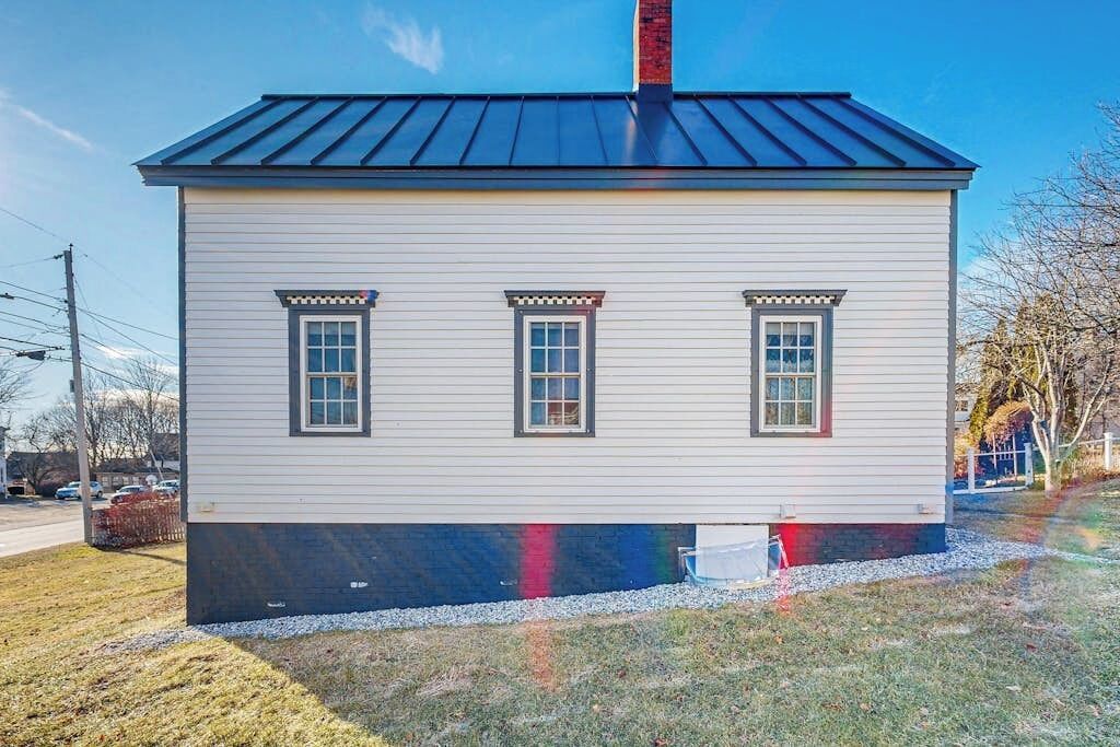 A small, single-story house with a dark roof, three windows with decorative trim, and a chimney.