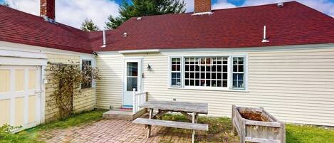 A single-story house with a red roof, a front door, a window with a grid pattern, a wooden bench, and a trash bin.