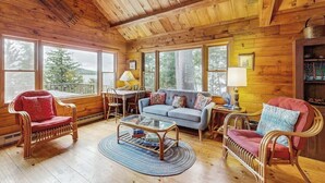 Un acogedor interior de cabaña con paredes de madera, un sofá, dos sillones, una mesa de centro y un escritorio con una lámpara.
