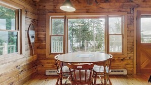 Interior de una cabaña de madera con una mesa redonda para comer, sillas de madera, ventanas amplias y una lámpara colgante.