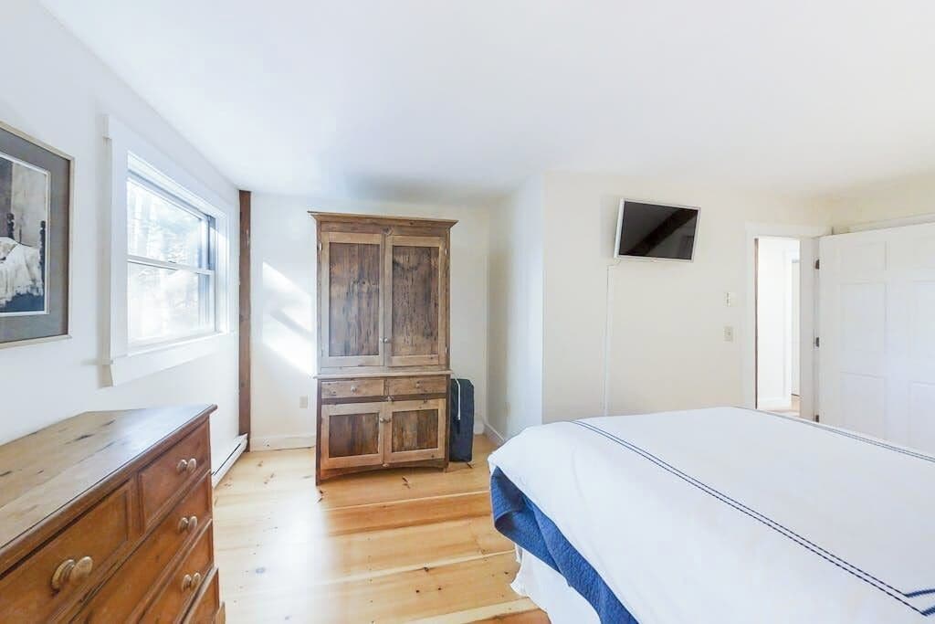 A bedroom with a wooden dresser, a bed with a white and blue bedspread, a television on the wall, and a large window.