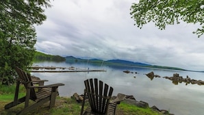 Duas cadeiras Adirondack perto de um lago tranquilo, com um cais e montanhas ao fundo.