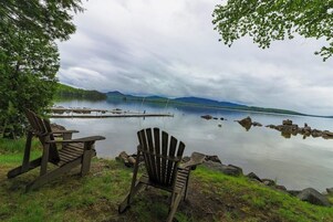 Dos sillas Adirondack frente a un lago tranquilo con un muelle y montañas al fondo.
