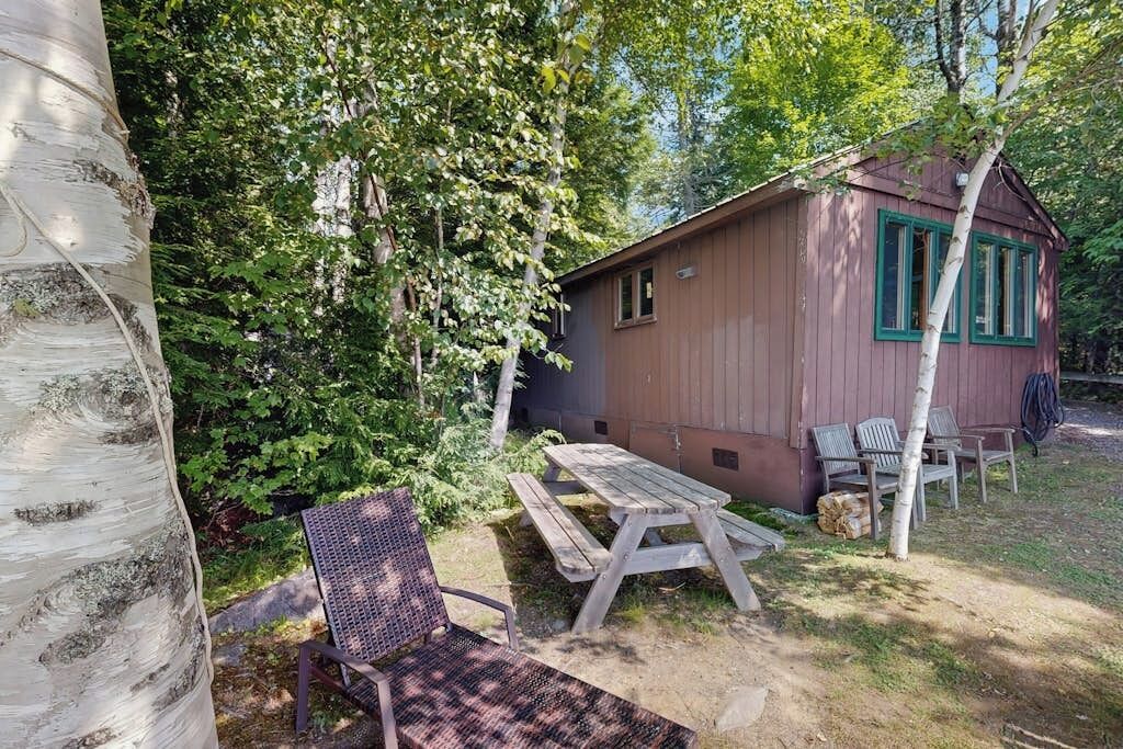 A wooden cabin with a picnic table and chairs, surrounded by trees.