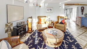 Ein Wohnzimmer mit einem großen Teppich mit Ankerprint, einem runden Couchtisch, zwei Sesseln, einem Vintage-Radio und einem an der Wand montierten Fernseher.