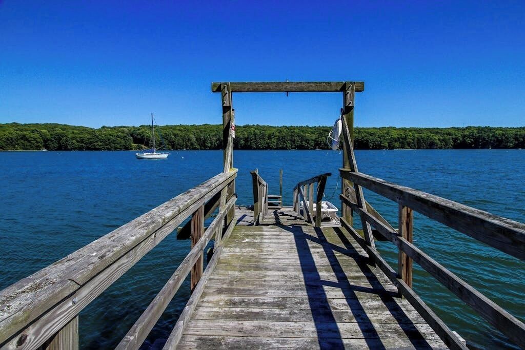 A wooden pier extending into a lake with a boat anchored in the distance and a forested shoreline.