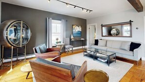 A modern living room with a round mirror, a sofa, a coffee table, and a large window. - Charming, Historic Cottage With Modern Upgrades - Stroll to Dining & Attractions (Boothbay Harbor)