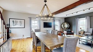 A dining area with a wooden table, chairs, a chandelier, and a large mirror. - Charming, Historic Cottage With Modern Upgrades - Stroll to Dining & Attractions (Boothbay Harbor)