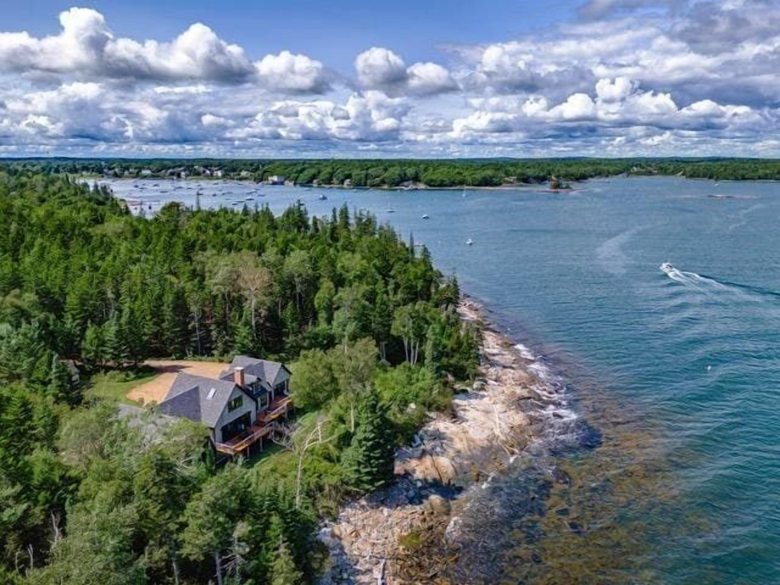 Une propriété située sur la côte, avec une maison entourée d’une forêt dense, offrant une vue sur une mer calme où naviguent des bateaux.