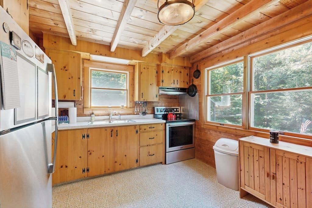 A wooden cabin kitchen with a refrigerator, stove, sink, and large windows.