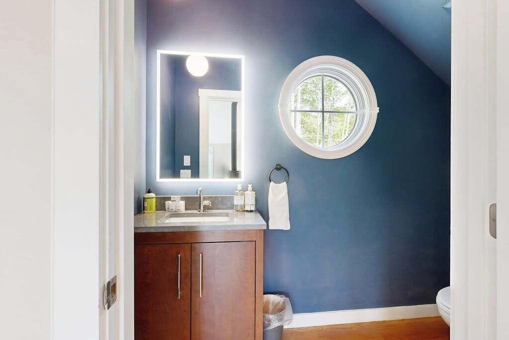 A bathroom with a dark blue wall, a round window, a mirror with a light, and a wooden cabinet.