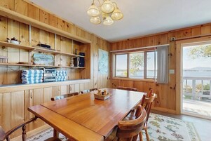 A wooden interior with a dining table, chairs, shelves with kitchen items, and a large window.
