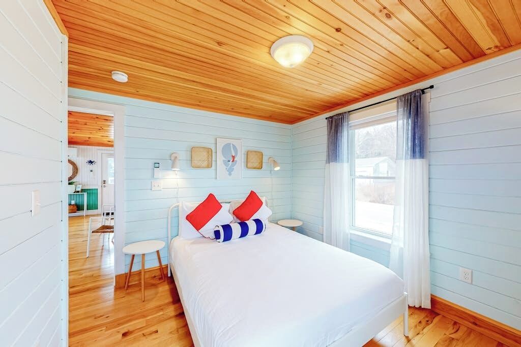 Un dormitorio con techo de madera, una cama con ropa blanca y almohadas rojas, una mesita pequeña y una ventana con cortinas.