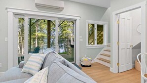 Un salon moderne avec un canapé gris, un escalier et une vue sur les arbres à travers des portes vitrées.