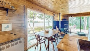 Une cabane en bois comprenant un coin repas, une cheminée et de grandes fenêtres donnant sur un paysage naturel.