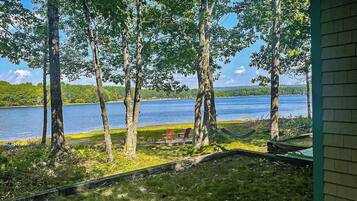 A lakeside view with trees, a hammock, and a deck with chairs.