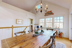 A dining area with a wooden table, chairs, a chandelier, and windows with a view.