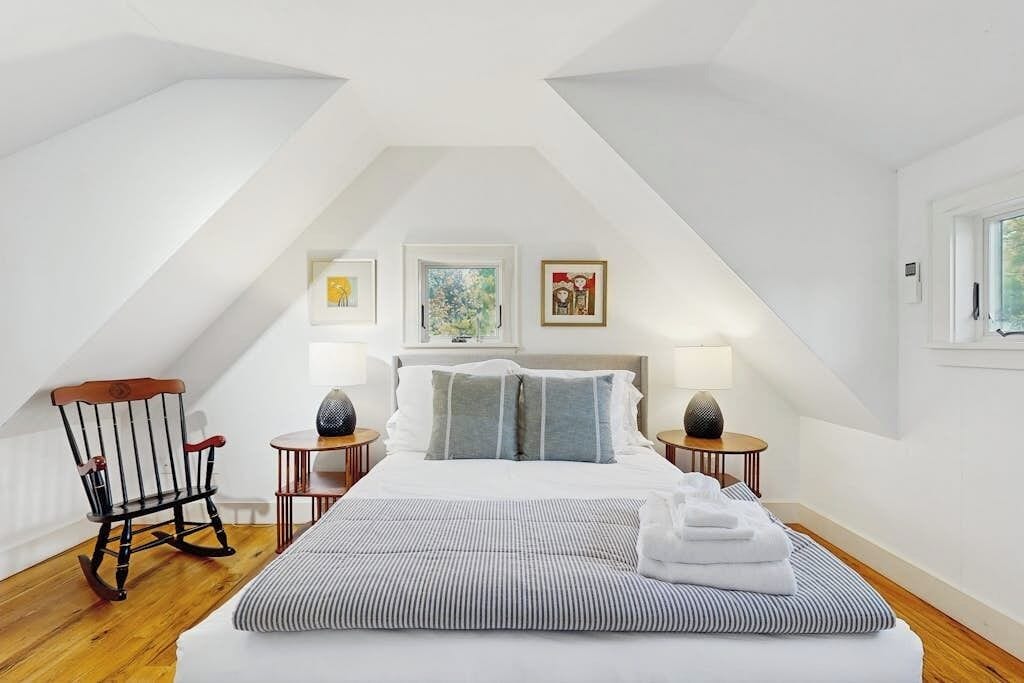 A bedroom with a sloped ceiling, a bed with pillows, two bedside tables with lamps, a rocking chair, and artwork on the walls.
