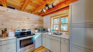 A log cabin kitchen with a stainless steel oven, microwave, and refrigerator, wooden ceiling beams, and a window with a view of trees. - Cozy Woodsy Cabin w/ a Deck, Firepit, Gas Grill, Free Wifi, & Full Kitchen (Greenville)