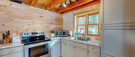 A log cabin kitchen with a stainless steel oven, microwave, and refrigerator, wooden ceiling beams, and a window with a view of trees.