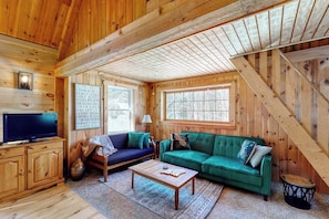 Interior de una cabaña de madera con una televisión, dos sofás, una mesa de centro y una escalera.