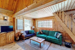 Interior de una cabaña de madera con una televisión, dos sofás, una mesa de centro y una escalera.