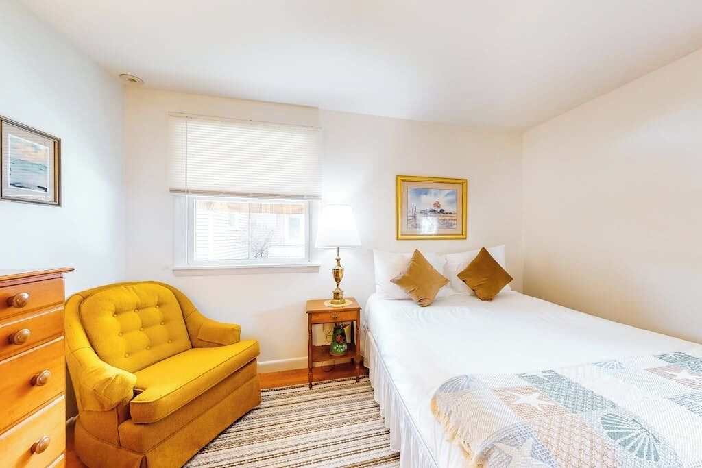 A bedroom with a bed, a yellow armchair, a dresser, a lamp, and a window with blinds.