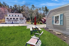 A grassy area with a picnic table and two Adirondack chairs, a white house with a garage, and a red mobile home. - Well-located, Dog-friendly Home - Walk to Footbridge & Barrett Park (Boothbay Harbor)