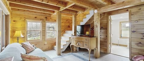 A wooden cabin interior with a sofa, a television, a staircase, and a view through the windows.