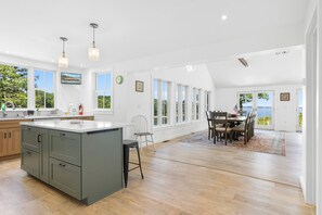 Ett rymligt kök med en stor köksö, en matplats med utsikt och ett vardagsrum med högt i tak och stora fönster.