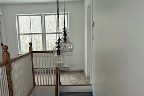 A staircase with a wooden railing, a white wall, and a window with a view of trees. - Gorgeous Oceanfront Home With Hot Tub, Modern Remodel, and Stunning Views (Wiscasset)