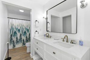 Un baño con un lavabo blanco, dos llaves, un espejo y una cortina de ducha con motivos forestales.