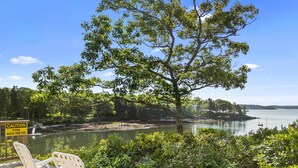 A wooden deck with two white chairs, overlooking a calm lake with boats, surrounded by lush greenery and trees. - Gorgeous Oceanfront Home With Hot Tub, Modern Remodel, and Stunning Views (Wiscasset)