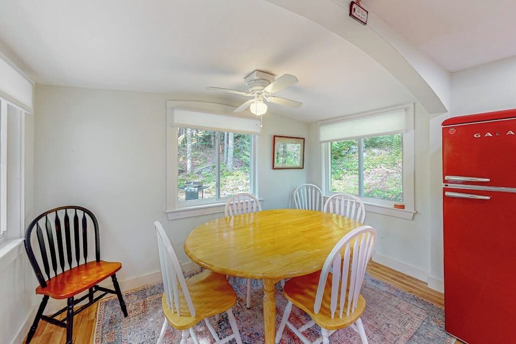Un coin repas avec une table ronde en bois, des chaises blanches, un réfrigérateur rouge et un ventilateur de plafond.