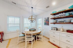 A dining area with a round table, white chairs, a chandelier, and a kitchen with white cabinets and shelves. - Perfectly Located Retreat With Grill, Fireplace, & Chef (Boothbay Harbor)