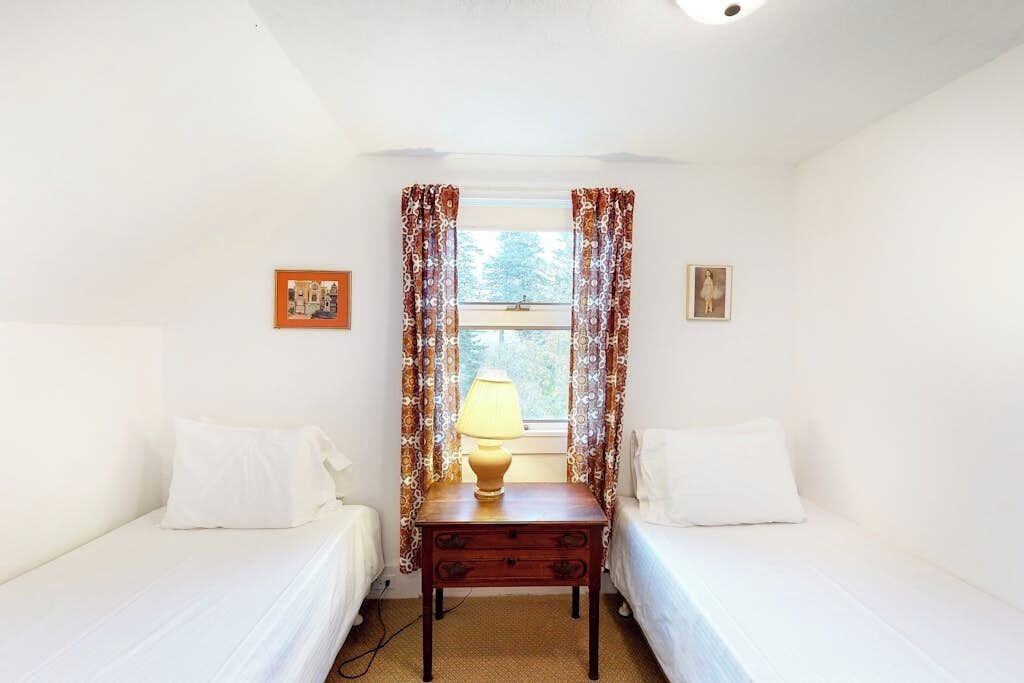 A room with two single beds, a small table with a lamp, and a window with patterned curtains.