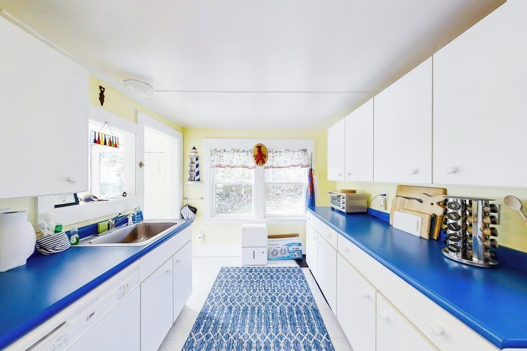 A kitchen with white cabinets, a blue countertop, a patterned rug, and a window with a view of trees.