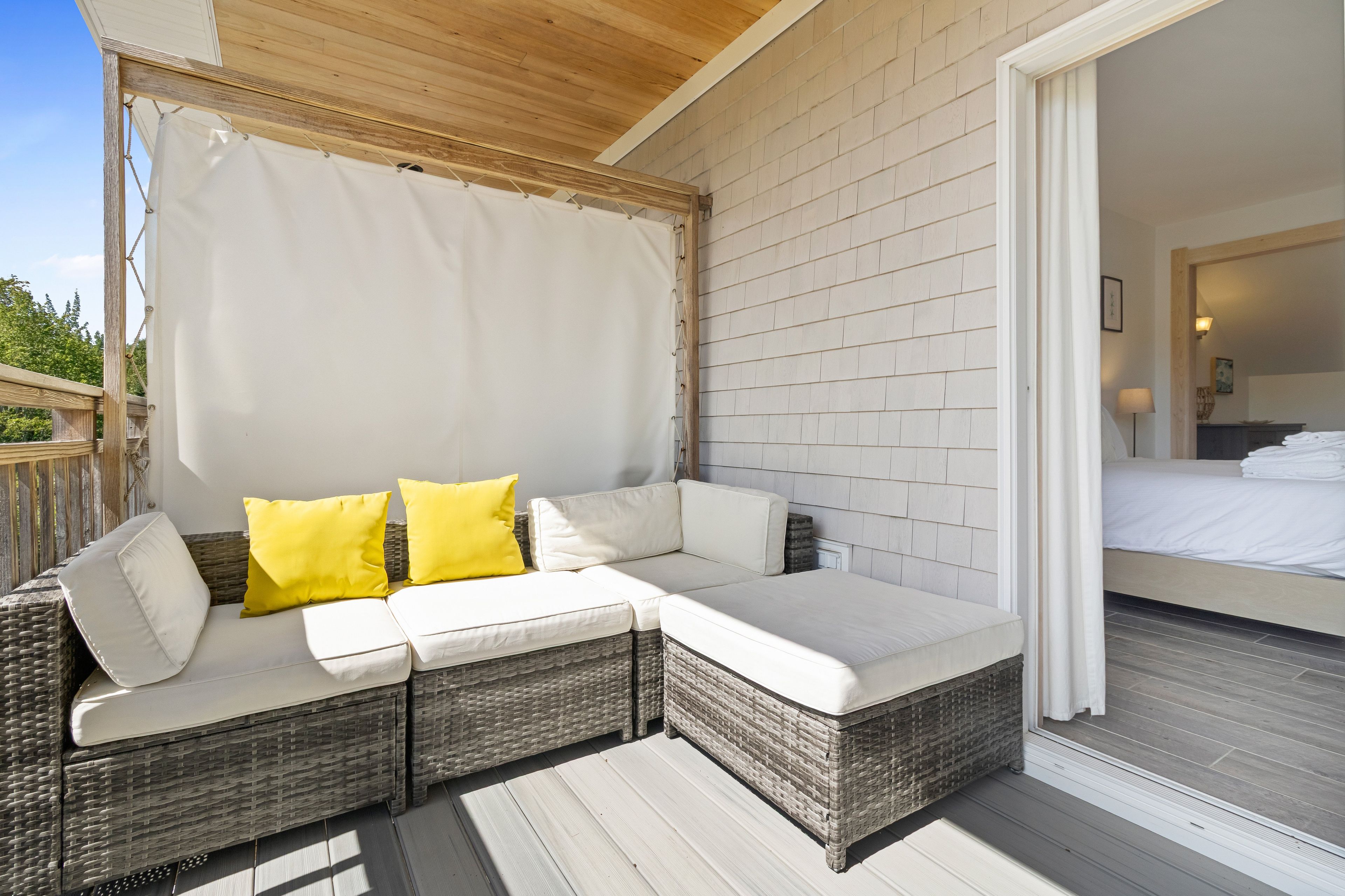 Een overdekt terras met een bank en kussens, een houten plafond en uitzicht op een slaapkamer met een bed en nachtkastje.