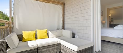 Un espacio de estar al aire libre techado con sofá y cojines, techo de madera, y vista a un dormitorio con cama y mesita de noche.