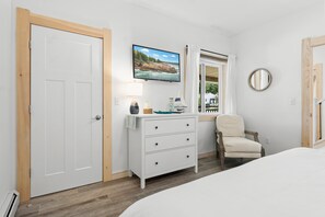 A bedroom with a white door, a dresser with drawers, a chair, a television on the wall, and a window with a view of the outdoors.