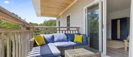 A covered outdoor seating area with a sofa and ottoman, a wooden deck, and a sliding glass door leading to an interior space.