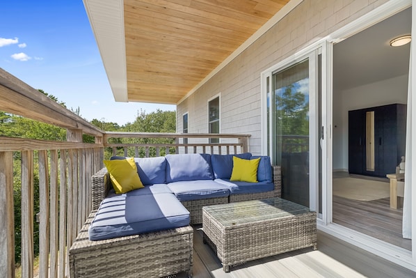 A covered outdoor seating area with a sofa and ottoman, a wooden deck, and a sliding glass door leading to an interior space. - Ocean Breeze (Owls Head)
