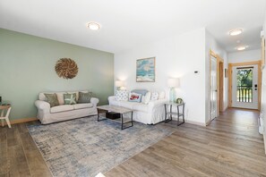 A living room with a white sofa, a coffee table, and a rug. There is a painting on the wall and a door leading outside.