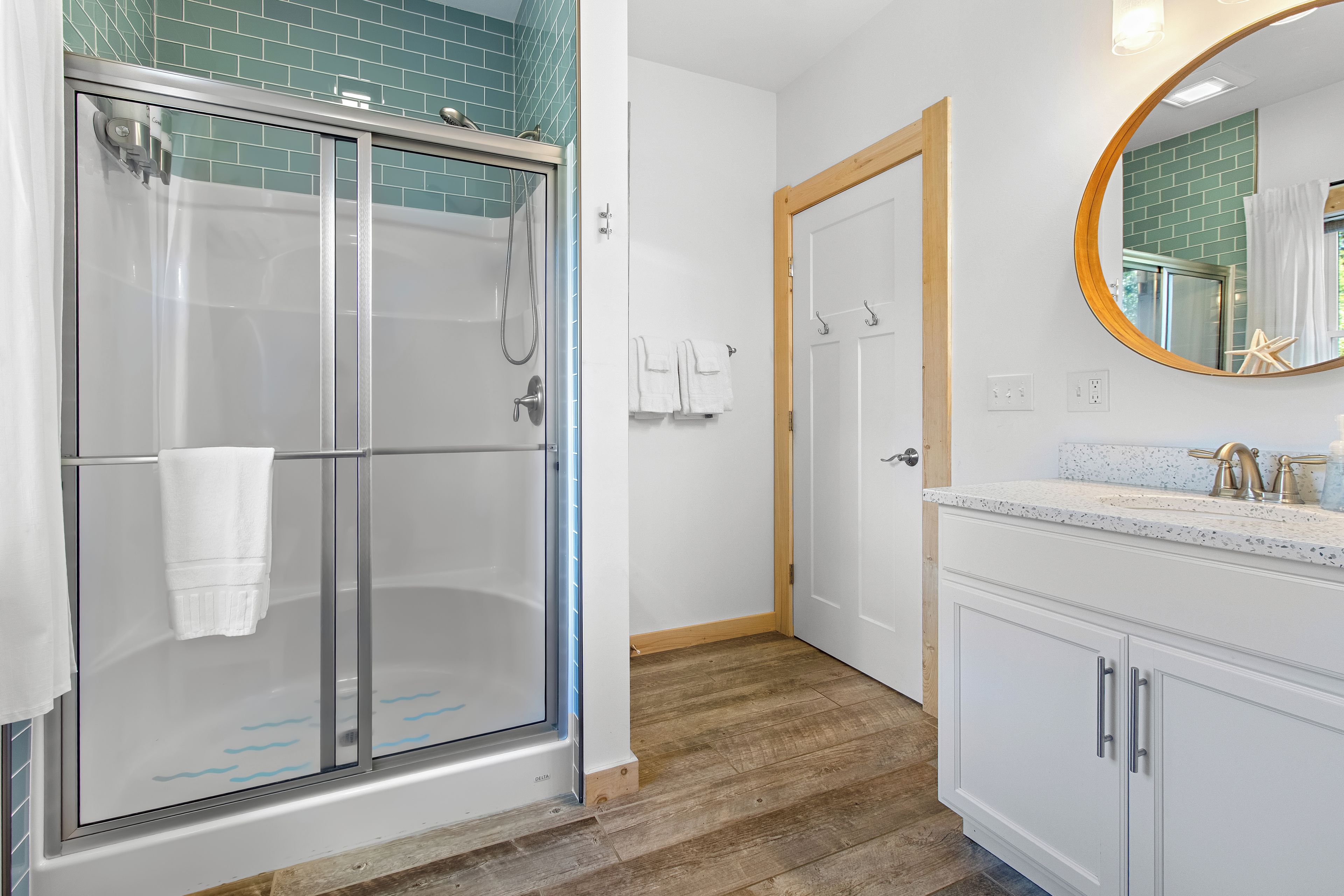 Een badkamer met een glazen douchecabine, een houten deur, een witte wastafel met een bekleurd aanrechtblad en een ronde spiegel.