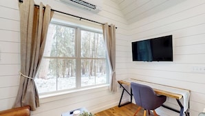 Ein Zimmer mit Holzboden, Schreibtisch mit Stuhl, Bett, Fernseher an der Wand und Fenster mit Vorhängen.