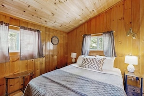 A wooden-paneled bedroom with a bed, two bedside tables, and curtains. - Quiet Lakefront Cottage With a Firepit, Private Beach, & two Kayaks (Ellsworth)