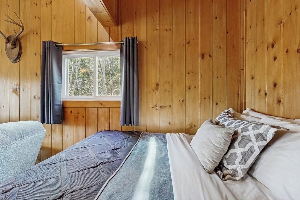 Une pièce de style cabane en bois, avec un lit, une fenêtre avec des rideaux et une tête d’animal empaillée accrochée au mur.