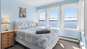 A bedroom with a bed, two bedside tables, a nightstand, a window with a view of the water, and a wall with a hanging decoration. - Cobbossee Hideaway, Lakefront Property With Dock for Swimming, Fishing, and Sunrises (Winthrop)